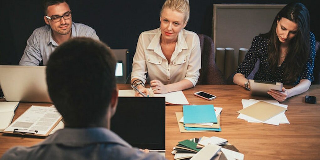 Homem de costas realizando apresentação com notebook a frente, diante de 3 pessoas