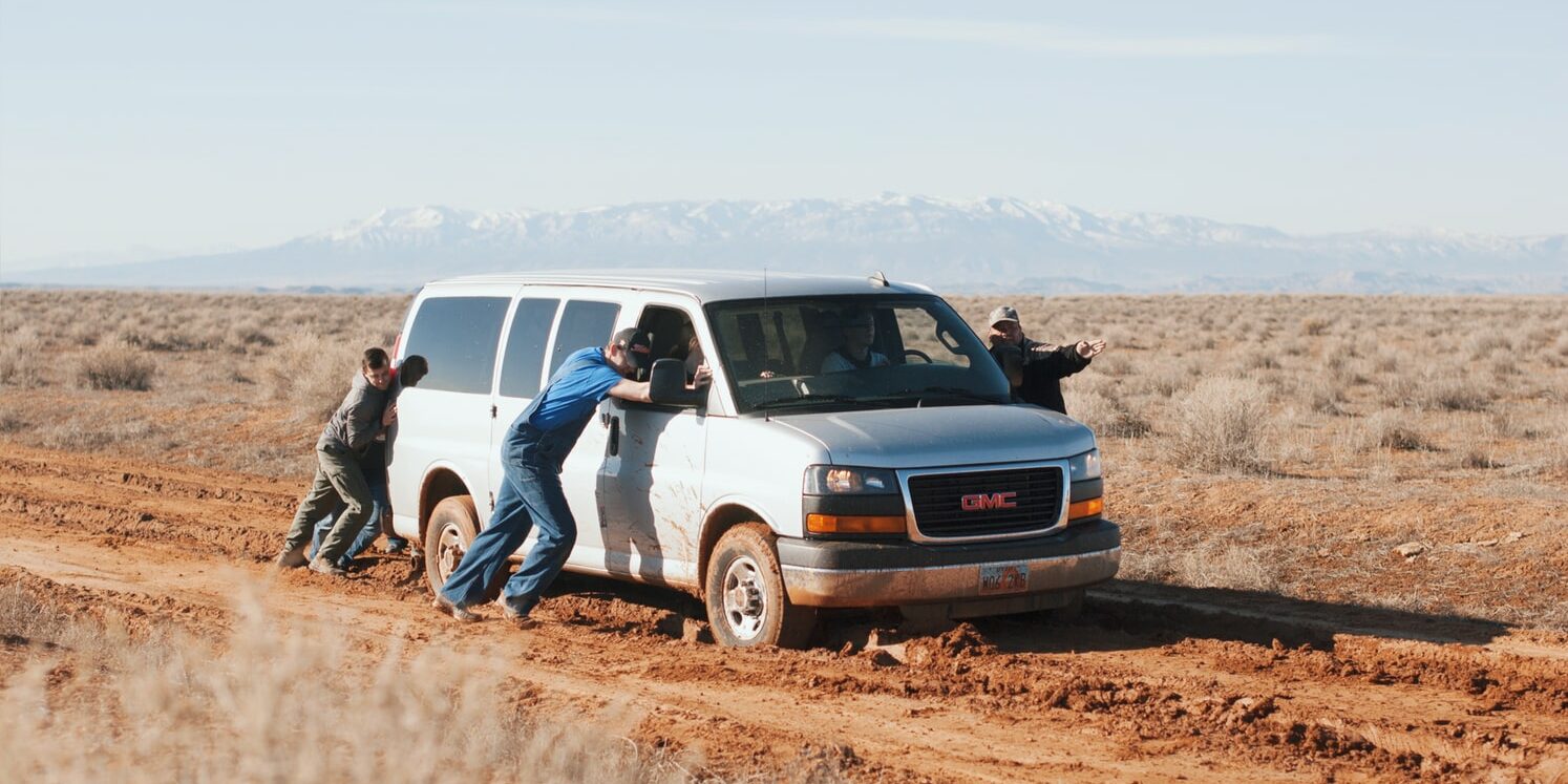 4 homens empurrando um carro num deserto. Um dos homens aponta para frente, orientando os demais